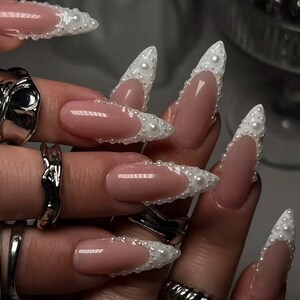 May include: Long, almond-shaped nails featuring a nude base color with white French tips adorned with small, raised white pearls. The fingers are decorated with several chunky, textured silver rings of varying shapes and sizes on multiple digits.