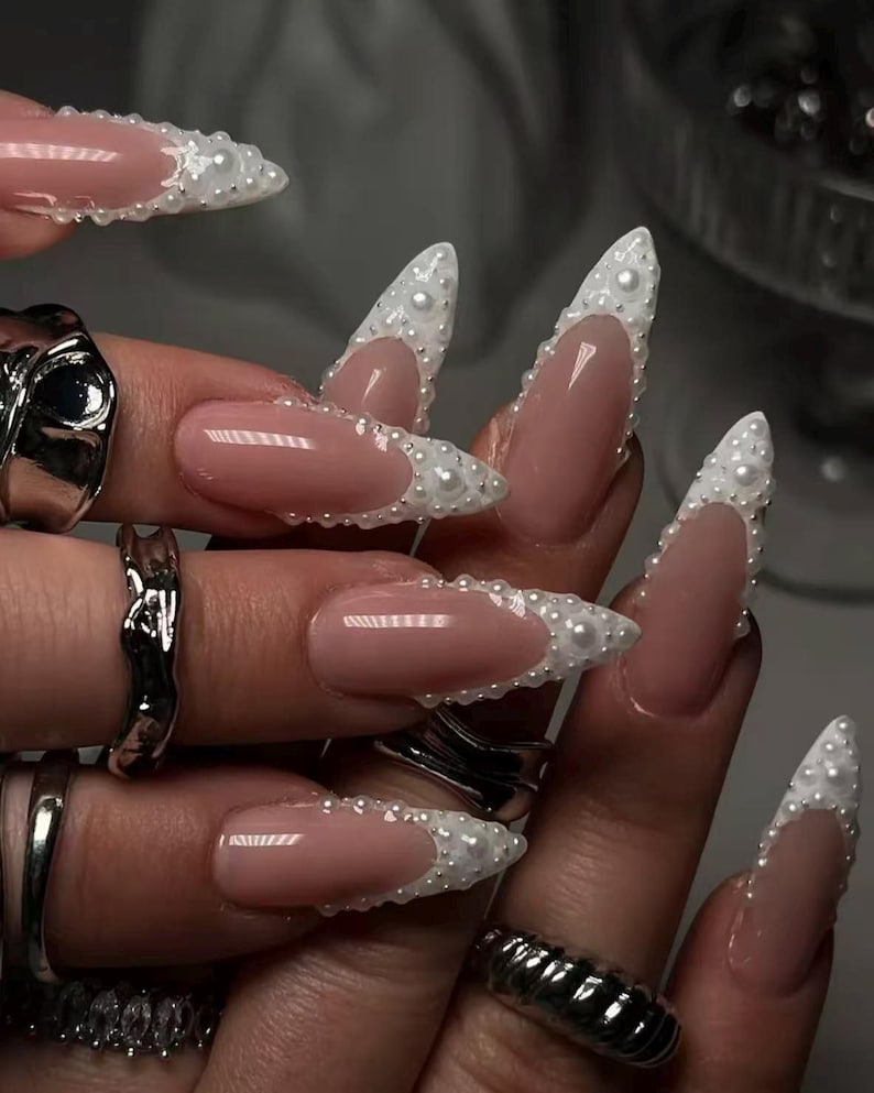 May include: Long, almond-shaped nails featuring a nude base color with white French tips adorned with small, raised white pearls. The fingers are decorated with several chunky, textured silver rings of varying shapes and sizes on multiple digits.