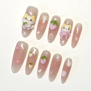 May include: Set of ten pink and white false nails with various designs. Some nails feature a small pink strawberry, a white paw print, a white heart, a white star, or a white swirl. Two nails feature a small 3D white cat holding a pink strawberry.