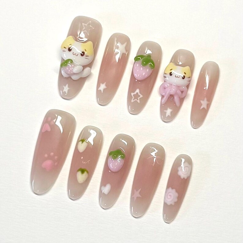 May include: Set of ten pink and white false nails with various designs. Some nails feature a small pink strawberry, a white paw print, a white heart, a white star, or a white swirl. Two nails feature a small 3D white cat holding a pink strawberry.