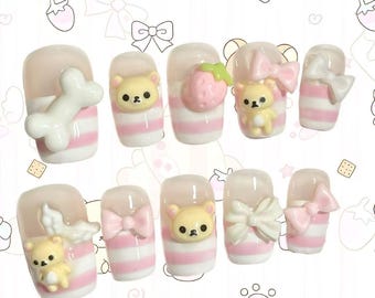 May include: A set of ten press-on nails with a light pink and white striped design. Each nail features 3D embellishments, including a bone, a strawberry, bows, and small yellow bear figures.