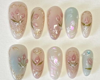 Könnte beinhalten: Set mit zehn falschen Nägeln in Pastellrosa und Blau. Jeder Nagel ist mit einem handgemalten Blumendesign verziert, das rosa Tulpen und goldene Akzente zeigt.