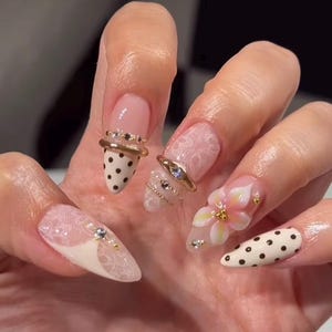 May include: Almond-shaped nails featuring a mix of designs including cream-colored polish with black polka dots, sheer white lace patterns, and a three-dimensional pink and white flower accent. The manicure is embellished with gold rings and small rhinestones.