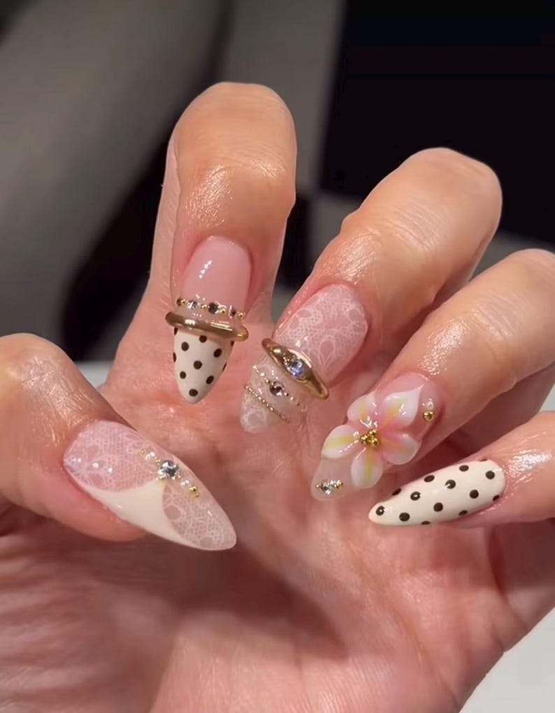 May include: Almond-shaped nails featuring a mix of designs including cream-colored polish with black polka dots, sheer white lace patterns, and a three-dimensional pink and white flower accent. The manicure is embellished with gold rings and small rhinestones.