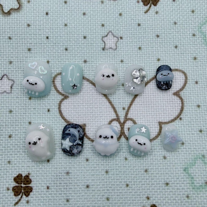 Peut inclure: Un ensemble de dix faux ongles avec divers motifs. Les ongles pr&eacute;sentent une palette de couleurs bleu clair, blanc et marine. Certains ongles comportent des personnages de dessins anim&eacute;s en 3D, des &eacute;toiles et un dauphin. D'autres ont des ornements argent&eacute;s. Les ongles sont dispos&eacute;s sur une surface &agrave; motifs bleu clair.