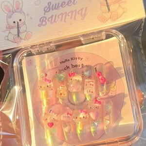 Peut inclure: Un ensemble de faux ongles transparents et iris&eacute;s avec des motifs Hello Kitty et des n&oelig;uds roses. Les ongles sont dans un &eacute;tui en plastique transparent avec le texte "Sweet Bunny" et une illustration de lapin.