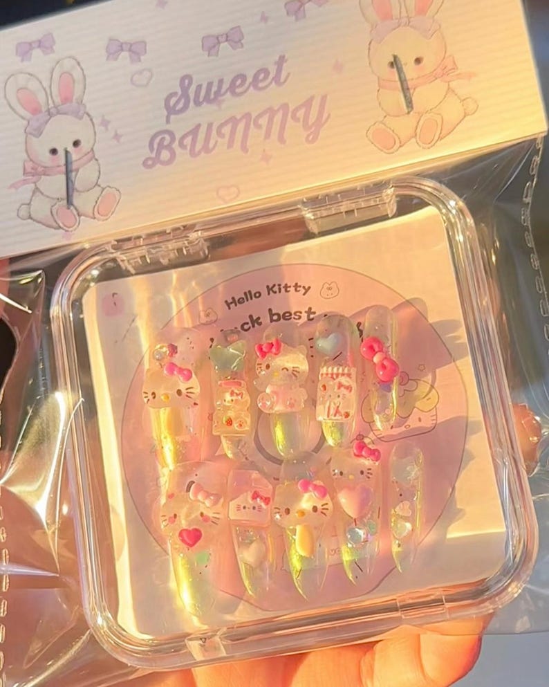 Peut inclure: Un ensemble de faux ongles transparents et iris&eacute;s avec des motifs Hello Kitty et des n&oelig;uds roses. Les ongles sont dans un &eacute;tui en plastique transparent avec le texte "Sweet Bunny" et une illustration de lapin.