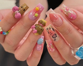 May include: Close-up of a hand with several different nail designs. The nails are decorated with pink, blue, and white colors, and feature 3D charms, including a cartoon cat, a seashell, and a bow. Some nails have a clear base with pink hearts and black musical notes.