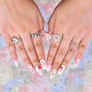 May include: A pair of hands displaying long, oval-shaped nails with intricate nail art. Designs include strawberries, bows, and floral patterns. Silver rings with heart, floral, and text details are also visible.