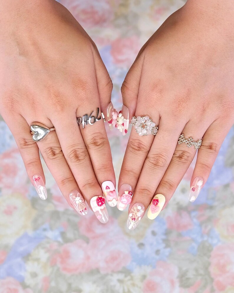May include: A pair of hands displaying long, oval-shaped nails with intricate nail art. Designs include strawberries, bows, and floral patterns. Silver rings with heart, floral, and text details are also visible.
