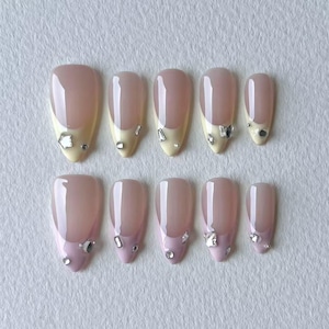 May include: A set of ten almond-shaped press-on nails featuring a nude base with French tips. The top row has pale yellow tips, and the bottom row has light pink tips. Each nail is decorated with small, clear, multifaceted rhinestones.