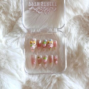 May include: A clear plastic case with 10 false nails. Each nail is decorated with a pink, white, and yellow cat design. The case has the text "Sweet 2 Teeth" printed on the top.