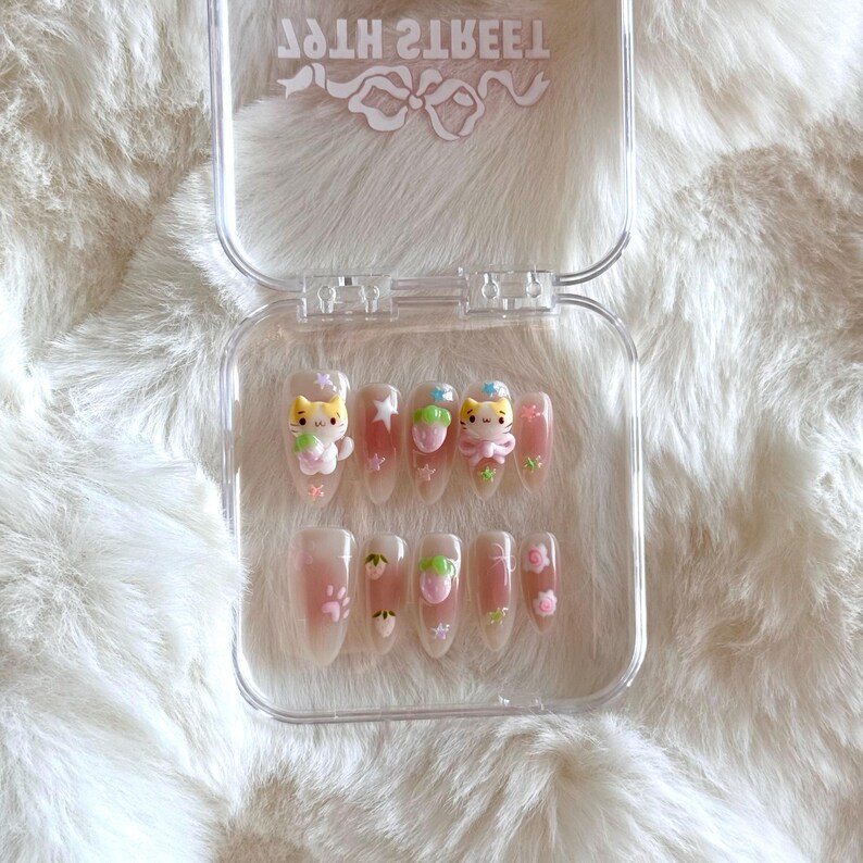 May include: A clear plastic case with 10 false nails. Each nail is decorated with a pink, white, and yellow cat design. The case has the text "Sweet 2 Teeth" printed on the top.