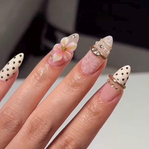 May include: Almond-shaped fingernails featuring diverse designs including cream-colored nails with black polka dots, a soft pink nail with a raised 3D floral accent, and nails adorned with white lace patterns, gold rings, and small embedded crystals.