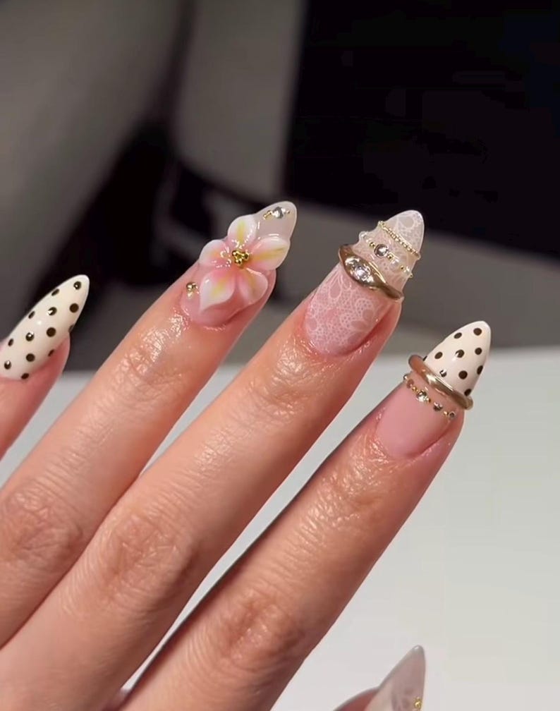 May include: Almond-shaped fingernails featuring diverse designs including cream-colored nails with black polka dots, a soft pink nail with a raised 3D floral accent, and nails adorned with white lace patterns, gold rings, and small embedded crystals.