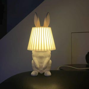 Op de afbeelding: Een witte konijnvormige lamp met een geplooide lampenkap. De lamp heeft rechtopstaande oren en een zittende konijnvorm. De lampenkap straalt een warme gloed uit, waardoor een gezellige sfeer ontstaat. Decoratieve verlichting.