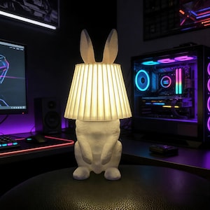 Op de afbeelding: Een witte lamp in de vorm van een konijn met een geplooide lampenkap. De lamp heeft lange oren en staat op een zwart oppervlak. De lamp is verlicht en geeft een warm licht. De achtergrond bevat een computeropstelling.
