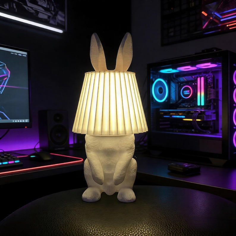 Op de afbeelding: Een witte lamp in de vorm van een konijn met een geplooide lampenkap. De lamp heeft lange oren en staat op een zwart oppervlak. De lamp is verlicht en geeft een warm licht. De achtergrond bevat een computeropstelling.