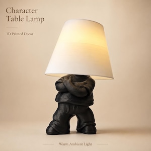 May include: A table lamp featuring a black figure with arms crossed, wearing a shirt and pants, topped with a white conical lampshade. The text reads: Character Table Lamp, 3D Printed Decor, Warm Ambient Light. The lamp emits a soft glow.