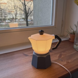Puede incluir: Una pequeña lámpara con forma de cafetera italiana. Tiene una base y un asa negras, y una sección superior crema iluminada. Está colocada sobre una superficie de madera cerca de una ventana. La lámpara tiene un cable blanco. Diseño italiano clásico.