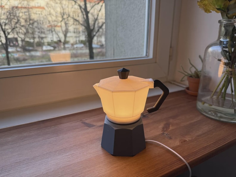 May include: A small, illuminated lamp shaped like a stovetop espresso maker. The lamp has a black base and handle, with a cream-colored, glowing upper section. It sits on a wooden surface near a window, with a white cord extending from the base.