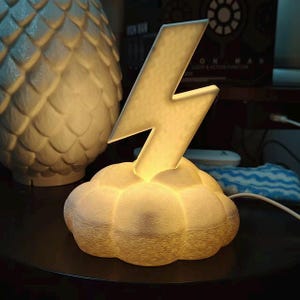 May include: A white, illuminated cloud-shaped lamp with a lightning bolt design. The lamp emits a warm glow, creating a soft ambiance. The lamp is on a dark surface, with another decorative object in the background.