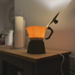 May include: A unique, illuminated coffee maker lamp. The lamp features a dark green base, an orange-colored top, and a black handle. The lamp is plugged into an electrical outlet with a white cord. The lamp is sitting on a wooden surface.