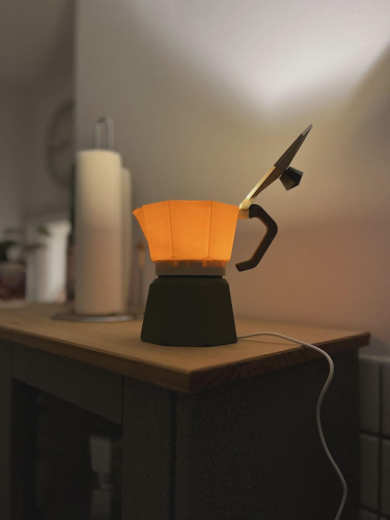 May include: A unique, illuminated coffee maker lamp. The lamp features a dark green base, an orange-colored top, and a black handle. The lamp is plugged into an electrical outlet with a white cord. The lamp is sitting on a wooden surface.