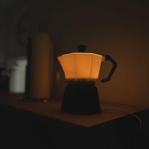 May include: A glowing, warm-toned coffee maker lamp with a black base and handle. The lamp is on a wooden surface, with a soft light emanating from within. The background is dark, creating a cozy, inviting atmosphere.