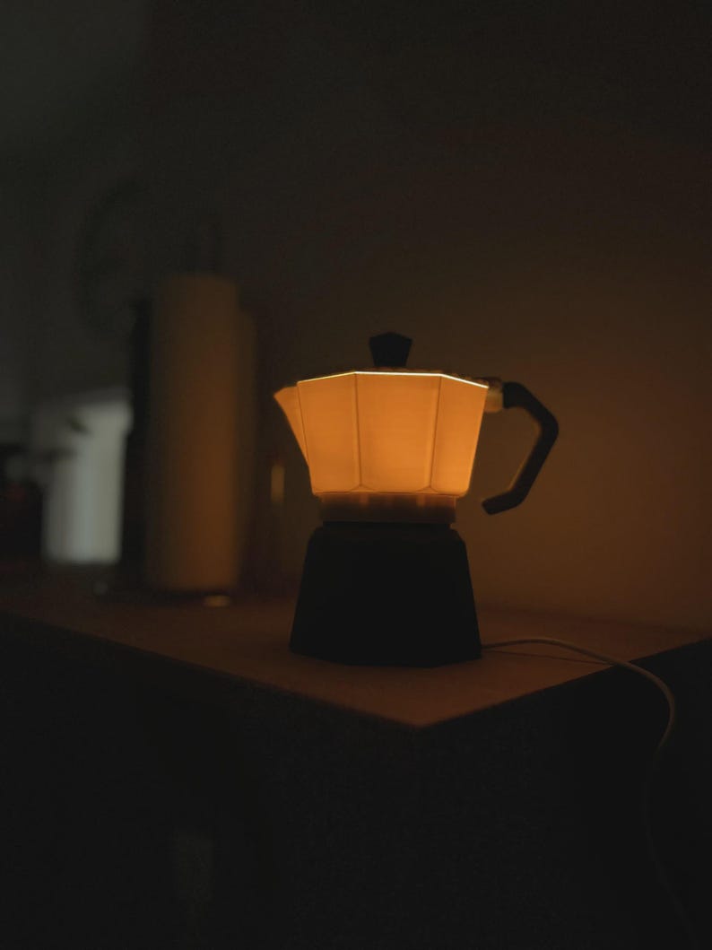 May include: A glowing, warm-toned coffee maker lamp with a black base and handle. The lamp is on a wooden surface, with a soft light emanating from within. The background is dark, creating a cozy, inviting atmosphere.