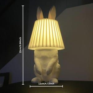 Op de afbeelding: Een witte tafellamp in de vorm van een konijn met een geplooide lampenkap. De lamp is 25 cm hoog en 12 cm breed. De oren van het konijn steken boven de lampenkap uit. De lamp is verlicht en geeft een warme gloed.