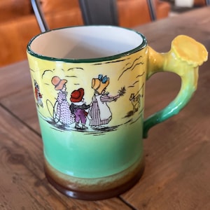 May include: A vintage ceramic mug with a whimsical illustration of children in a field. The mug features a yellow and green color scheme with a brown base and a unique handle shaped like a tree branch. The mug is likely a collectible item.