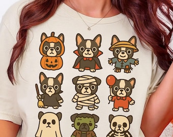 Puede incluir: Camiseta de color crema suave con una cuadrícula de perros de dibujos animados disfrazados para Halloween. Los perros llevan disfraces de calabaza, vampiro, momia y fantasma. La camiseta tiene la inscripción "Soft Cream".