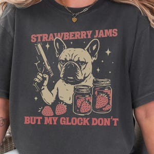 以下が含まれることがあります： フレンチブルドッグが銃を持ち、イチゴジャムの瓶と「STRAWBERRY JAMS BUT MY GLOCK DON'T」の文字が描かれた、濃いグレーのTシャツ。「PEPPER」の文字が左上にあります。