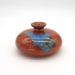 May include: A small, ceramic vase with a rounded body and a narrow opening. The vase is primarily a reddish-brown color, with blue and gray abstract designs. The vase is approximately 4 inches in diameter and 3 inches tall.