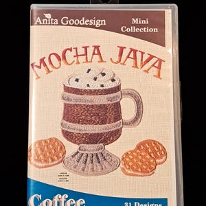 May include: A clear plastic case containing an Anita Goodesign Mini Collection embroidery pattern set titled Mocha Java. The cover features an embroidered coffee mug with whipped cream and two cookies, with the text Coffee and 31 Designs.