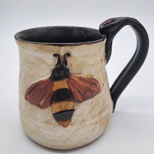 May include: A ceramic mug with a cream-colored exterior and a dark brown interior. The mug features a raised, sculpted bee design on the front with brown and gold wings and a striped body. It has a thick, dark brown handle attached to the side.