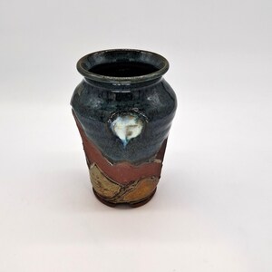 May include: A handcrafted ceramic vase with a dark blue glazed top and a textured, layered design in shades of red, brown, and gold. The vase features a unique, organic shape and a white accent near the center.