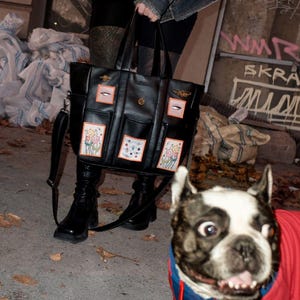 May include: A black leather tote bag with multiple pockets and embroidered floral designs. The bag has a long strap and is worn by a person with a black and white dog.