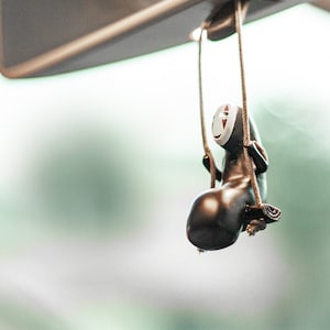 May include: A black and white No Face character from the movie Spirited Away hanging from a rearview mirror in a car. The character is on a swing and has a red and white face.