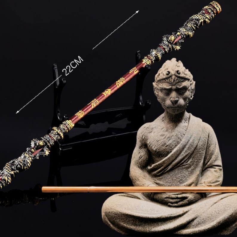 K&ouml;nnte beinhalten: Ein roter und goldener Stab mit einem Drachenkopf an der Spitze und chinesischen Schriftzeichen entlang des Schaftes. Der Stab ist 56 cm lang und wird auf einem schwarzen St&auml;nder pr&auml;sentiert. Eine Steinstatue eines Affen in meditativer Pose befindet sich im Vordergrund.