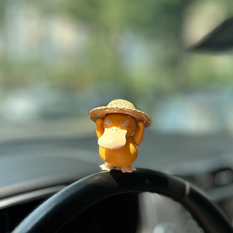 May include: A yellow duck figurine wearing a straw hat, with a grumpy expression, is perched on a black steering wheel.