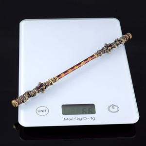 K&ouml;nnte beinhalten: Ein goldener und schwarzer dekorativer Stab mit filigranen Details und chinesischen Schriftzeichen. Der Stab liegt auf einer wei&szlig;en digitalen Waage, die 6,6 Gramm anzeigt.