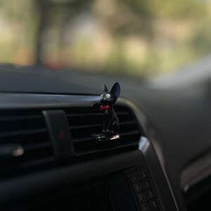 May include: Black cat figurine with a red bow tie, sitting on a car air vent.