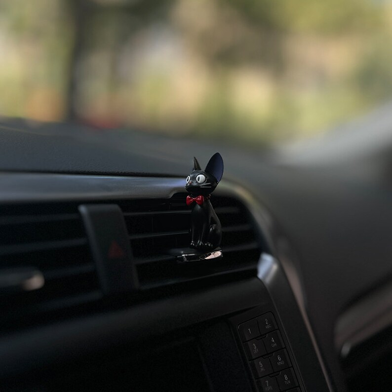 May include: Black cat figurine with a red bow tie, sitting on a car air vent.