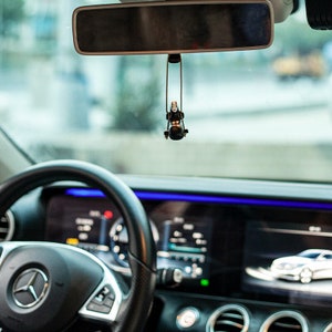 May include: A black car interior with a steering wheel, dashboard, and a rearview mirror. A small black and white character figurine hangs from the rearview mirror.