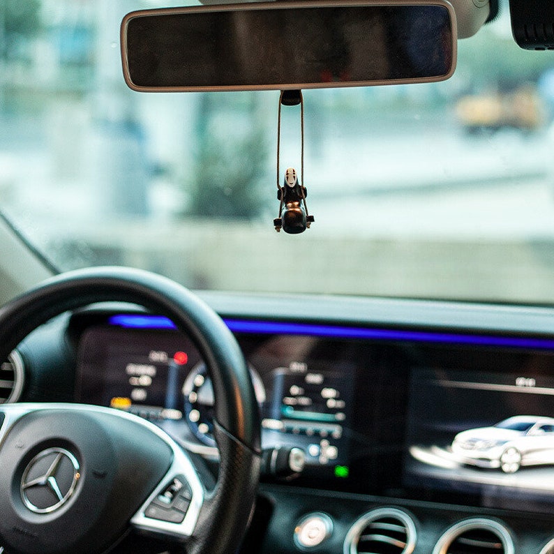 May include: A black car interior with a steering wheel, dashboard, and a rearview mirror. A small black and white character figurine hangs from the rearview mirror.