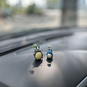 May include: Two small figurines of characters from the animated film My Neighbor Totoro. The figurines are gray and blue and are holding small green leaves. The figurines are sitting on a black surface.