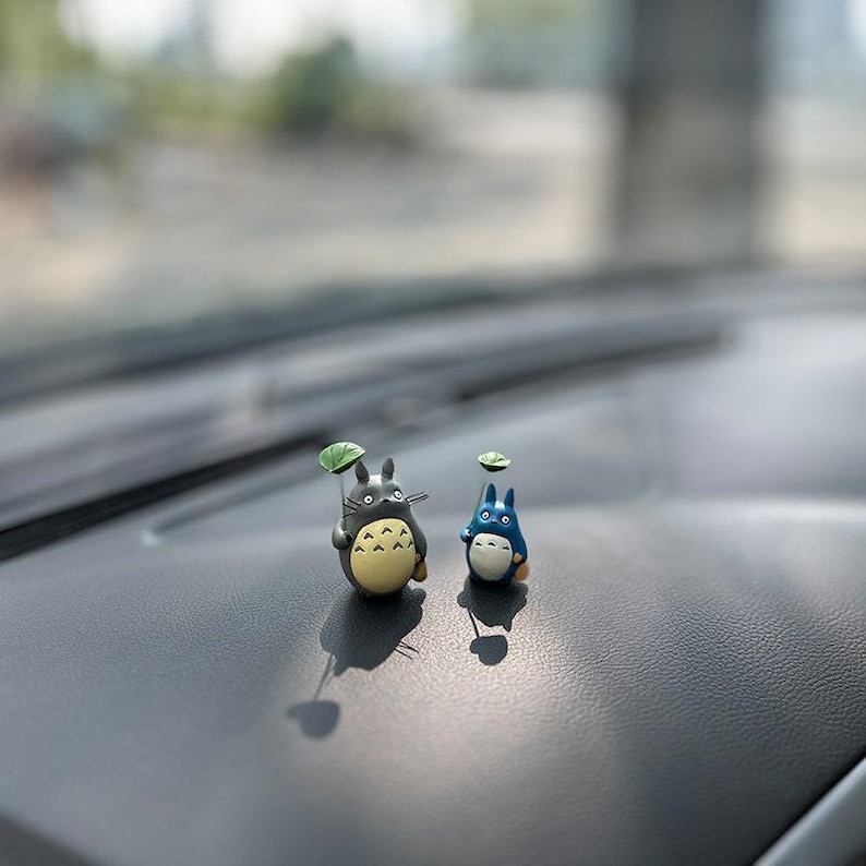 May include: Two small figurines of characters from the animated film My Neighbor Totoro. The figurines are gray and blue and are holding small green leaves. The figurines are sitting on a black surface.