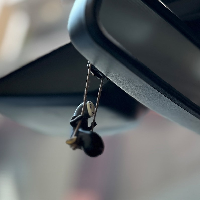 May include: A black and white character figurine hanging from a rearview mirror in a car. The character is wearing a mask and is holding onto a rope.